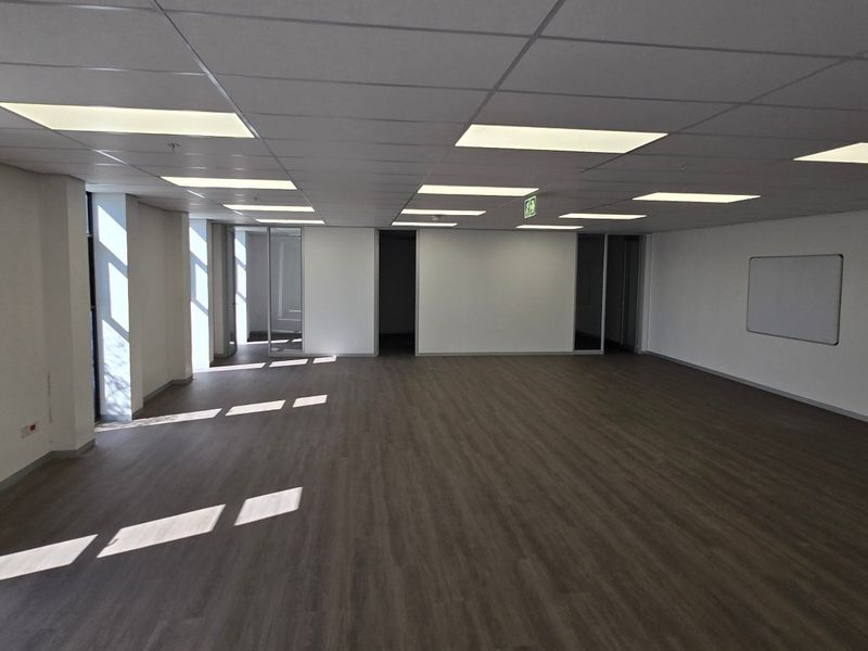 A spacious, empty open-plan office area featuring modern laminate flooring, a suspended ceiling with recessed lighting, and large windows providing natural light.