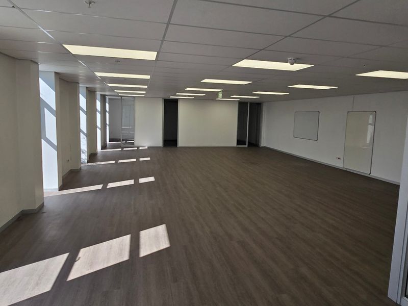 A spacious, vacant open-plan office area featuring modern laminate flooring, a suspended ceiling with recessed lighting, and large windows providing natural light.