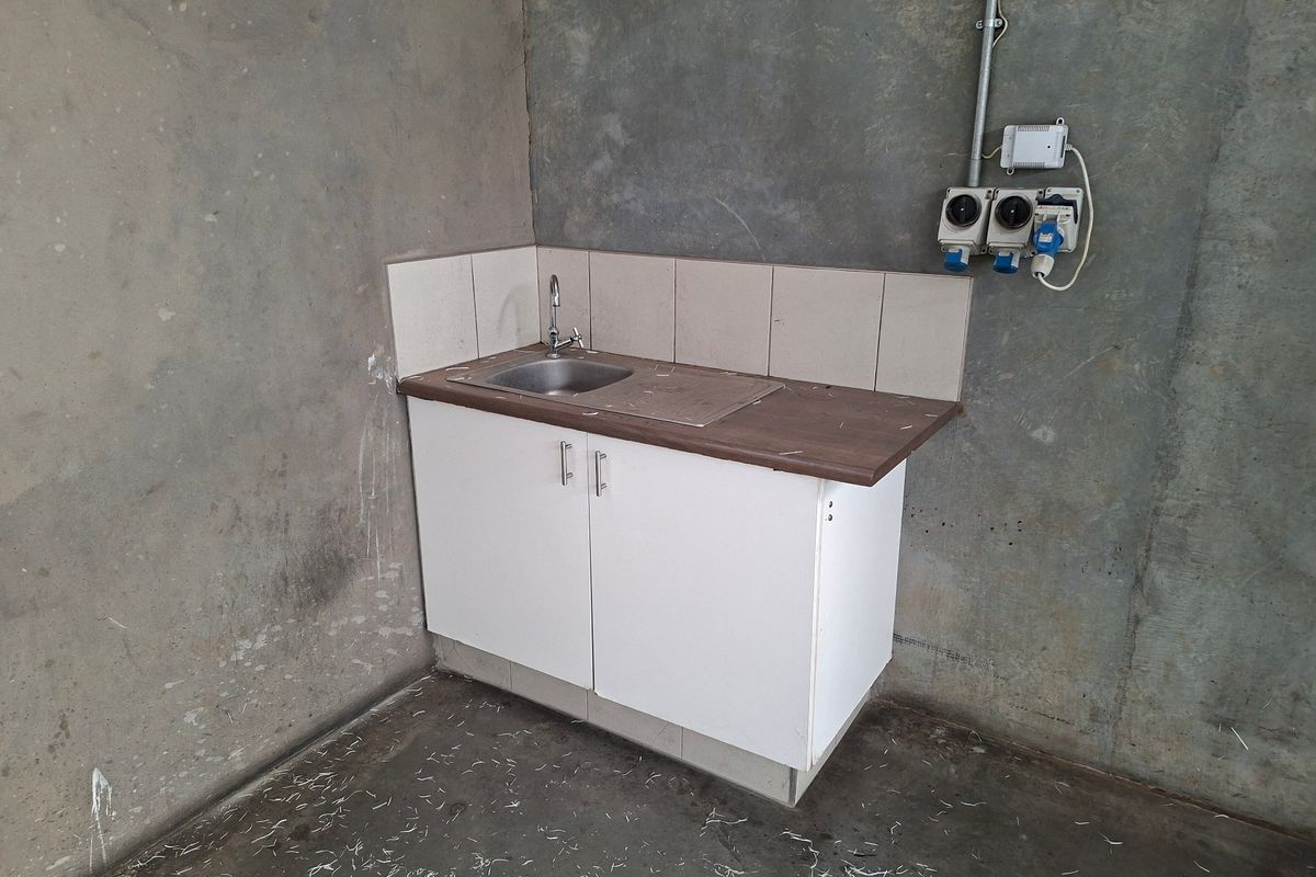 A basic utility area featuring a small sink with base cabinetry, tiled splashback, and exposed industrial electrical fittings against unfinished concrete walls.