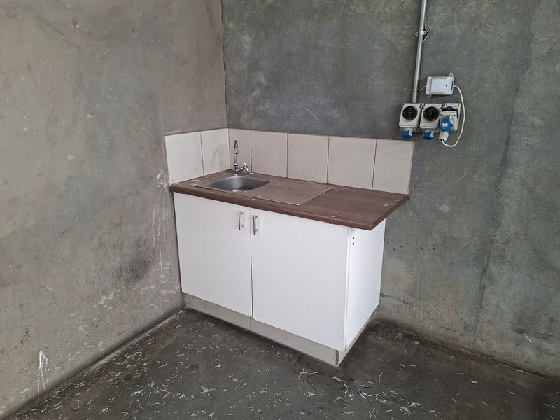 A basic utility area featuring a small sink with base cabinetry, tiled splashback, and exposed industrial electrical fittings against unfinished concrete walls.