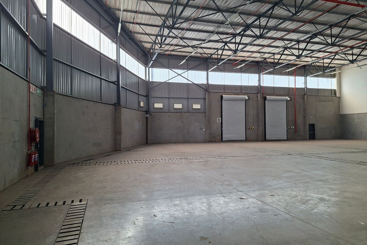 Spacious industrial warehouse featuring high ceilings with exposed steel trusses, concrete flooring, roller shutter doors, and integrated fire safety systems.