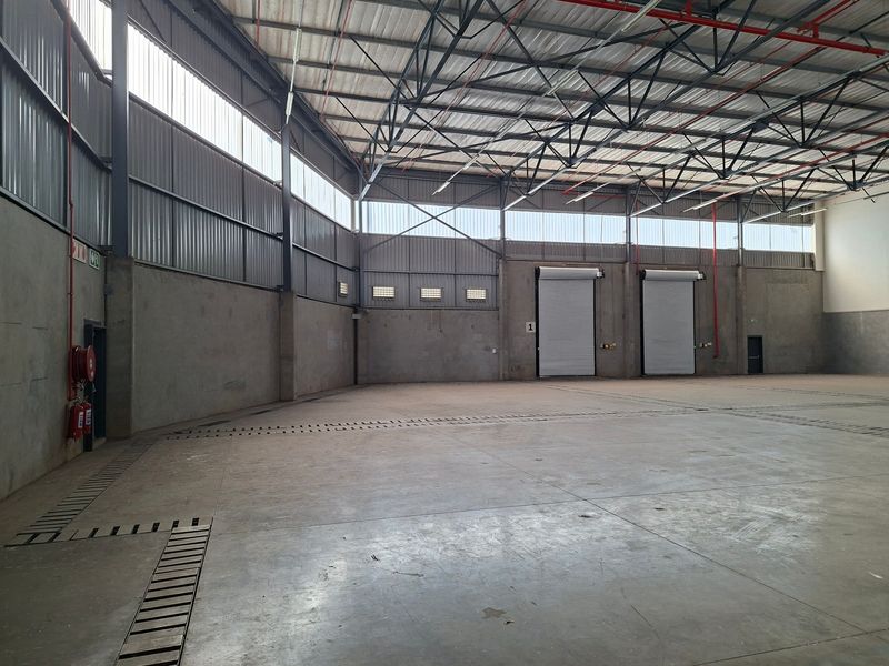 Spacious industrial warehouse featuring high ceilings with exposed steel trusses, concrete flooring, roller shutter doors, and integrated fire safety systems.