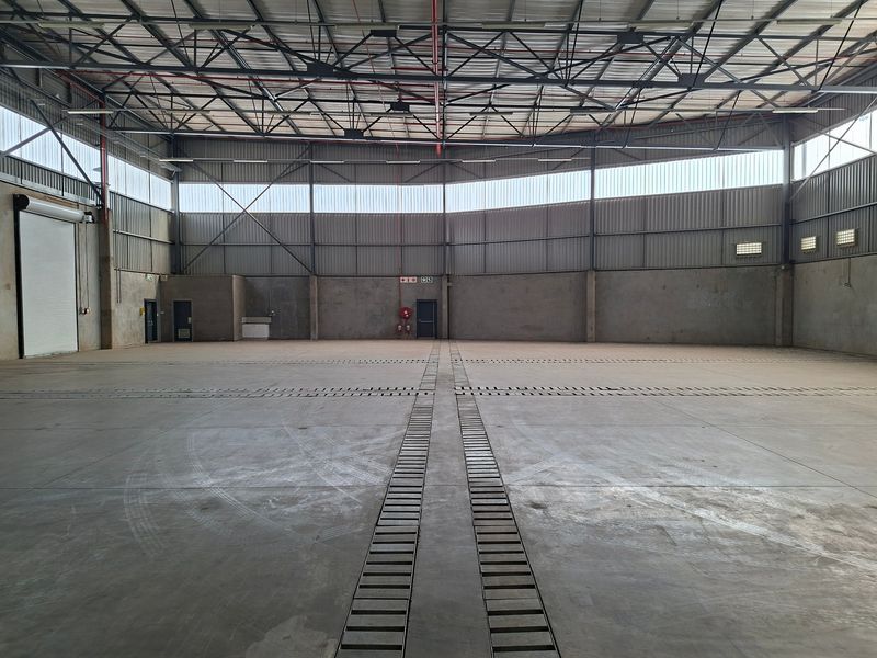 Spacious industrial warehouse featuring high ceilings with exposed steel trusses, concrete flooring with drainage channels, and a large roller shutter door for easy access.