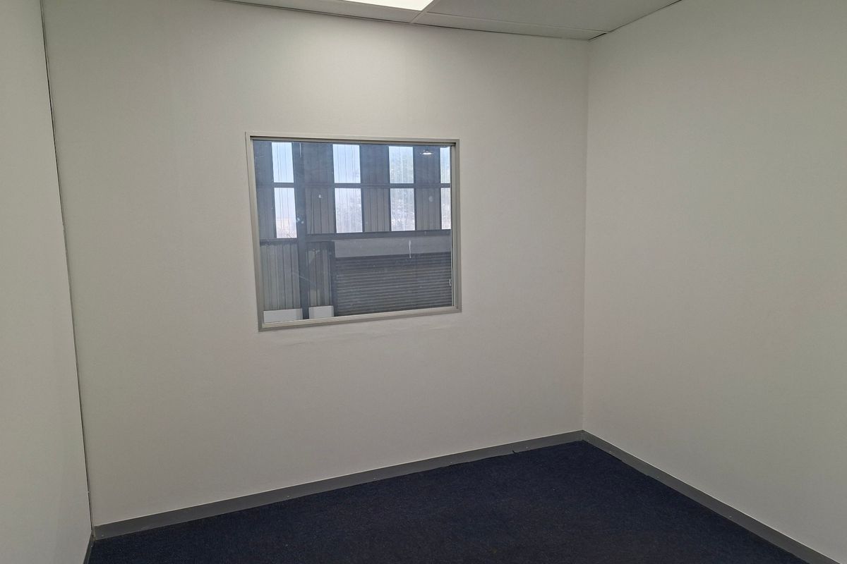 A clean, empty office space featuring neutral walls, carpeted flooring, and an internal window looking out into the industrial warehouse area.