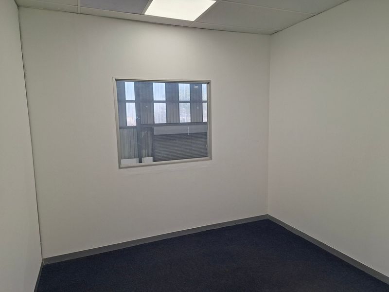 A clean, empty office space featuring neutral walls, carpeted flooring, and an internal window looking out into the industrial warehouse area.