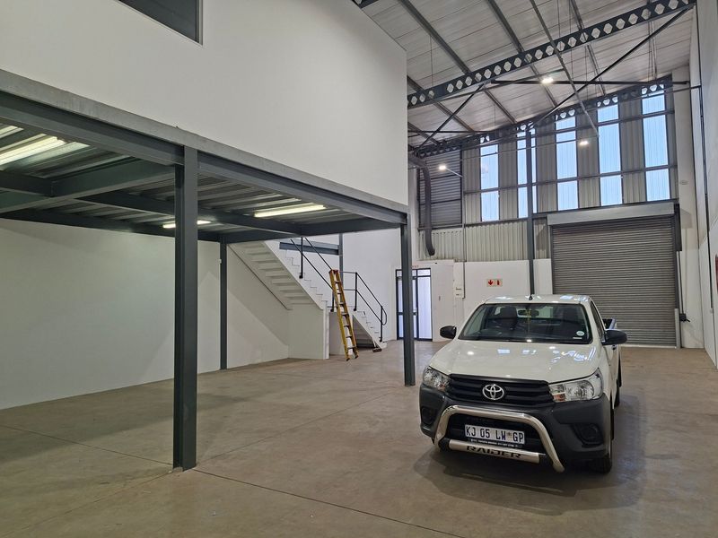 Spacious industrial warehouse unit featuring a steel mezzanine level, concrete flooring, high ceilings with exposed trusses, and a large roller shutter door.