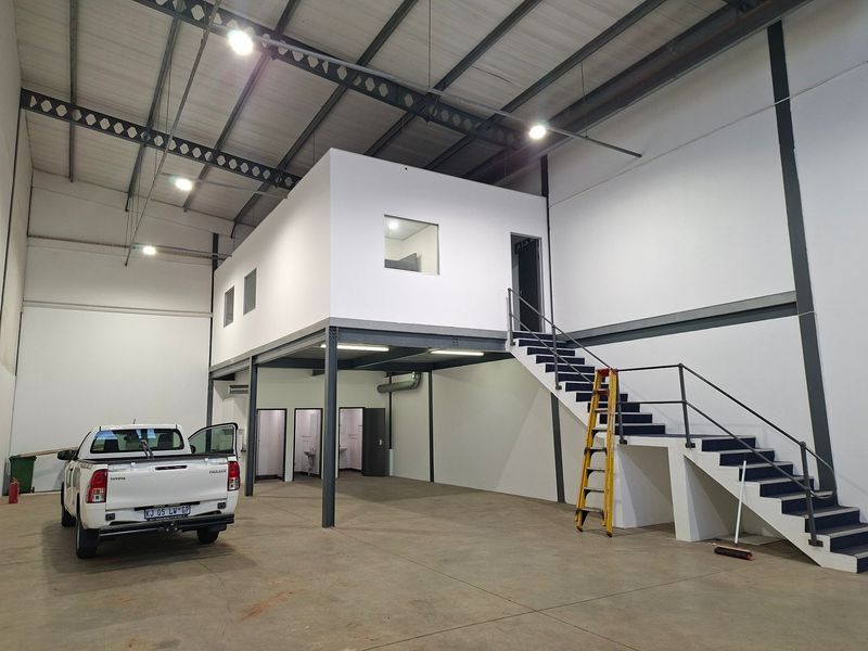 Spacious industrial warehouse featuring a mezzanine level, steel staircase, concrete flooring, and integrated office or utility rooms with high-clearance ceilings.