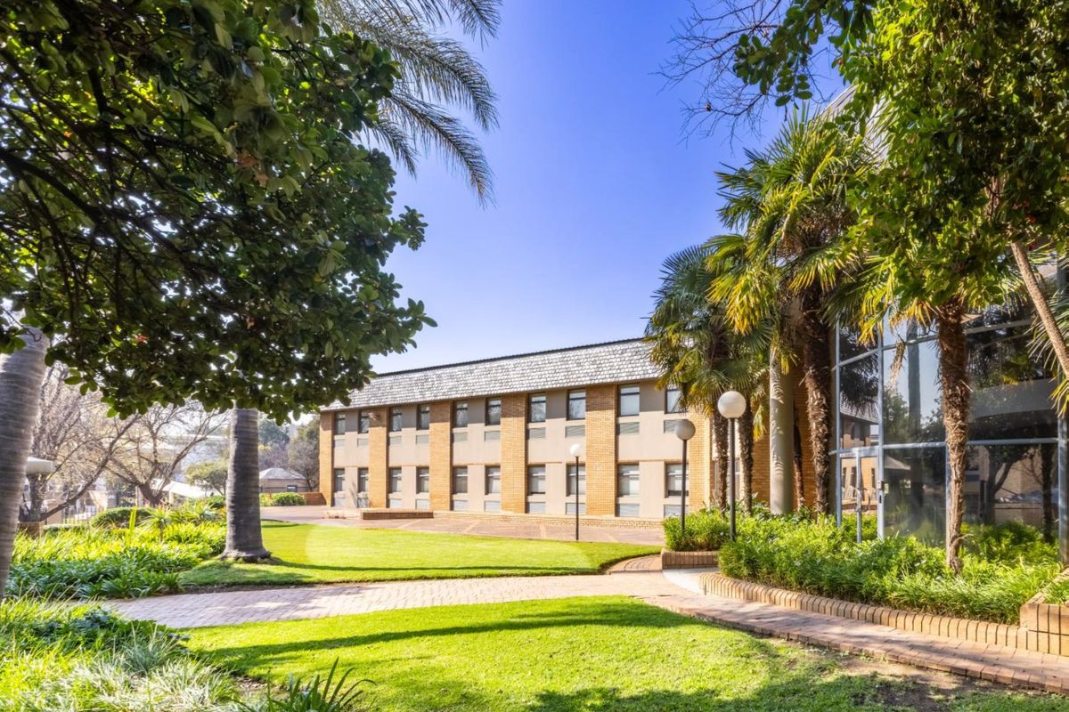 A well-maintained commercial building exterior featuring a brick facade, large glass windows, and landscaped garden areas with paved walkways.