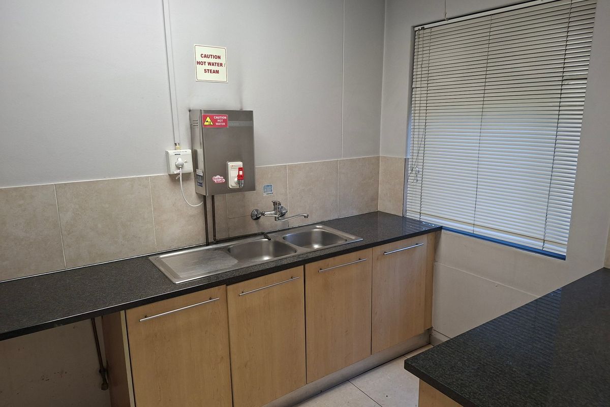 A functional commercial kitchenette featuring a stainless steel double sink, boiling water dispenser, laminate cabinetry, and tiled splashback with natural light from a window.