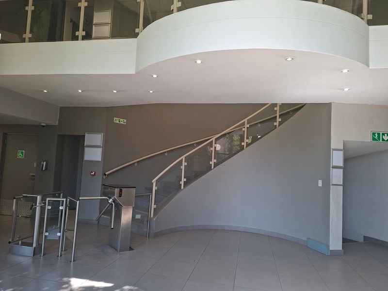 Modern commercial building entrance lobby featuring a curved staircase with glass balustrades, tiled flooring, and integrated turnstile access control systems.