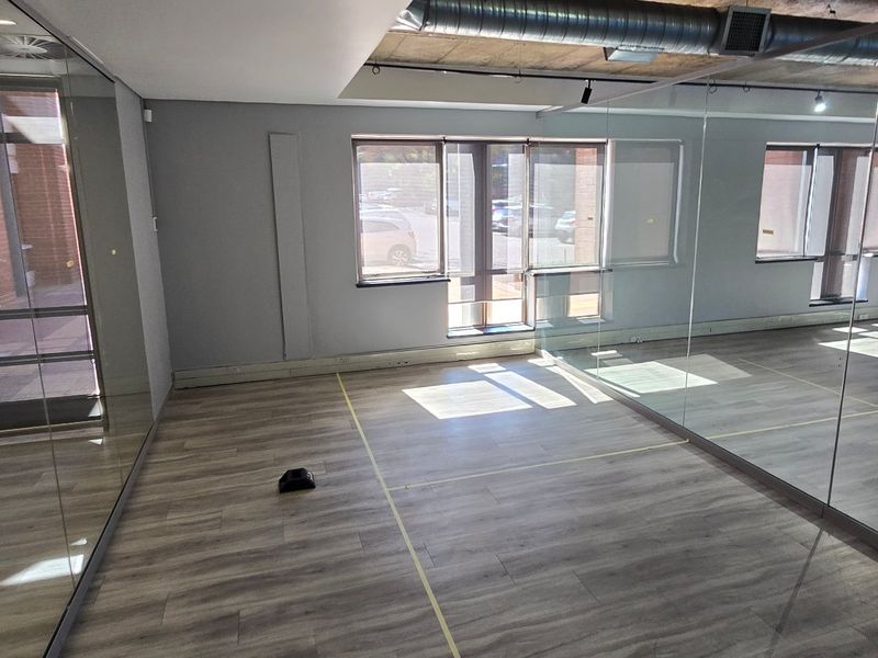 A modern, open-plan commercial studio space featuring full-length wall mirrors, exposed industrial ceilings, and durable laminate wood flooring with large glass partitions.