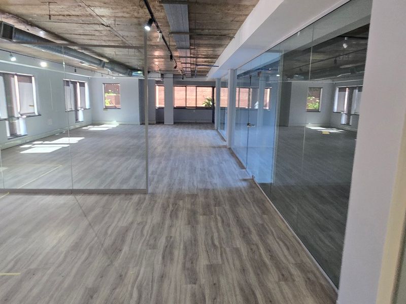 Modern open-plan office space featuring exposed concrete ceilings, industrial ducting, glass partitioning, and durable laminate flooring, offering a bright and professional work environment.