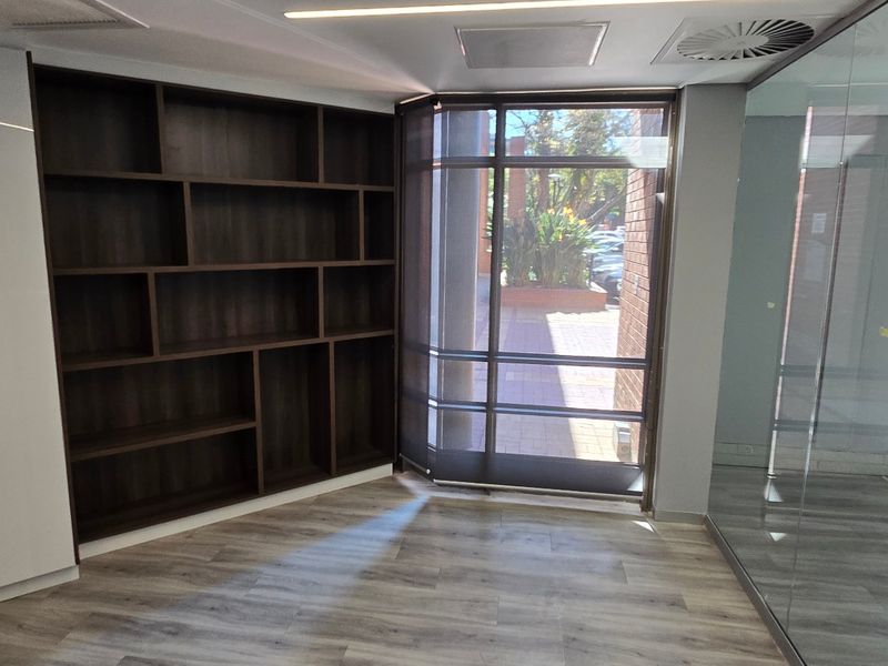 Modern office space featuring built-in dark wood shelving, glass partition walls, and light-toned laminate flooring with ample natural light.