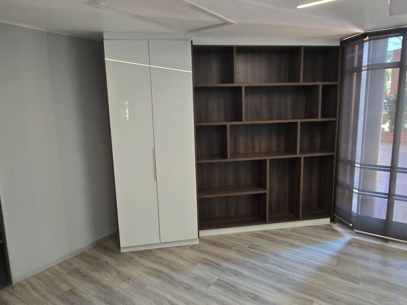 A modern, bright office space featuring built-in dark wood shelving, a white storage cupboard, and large windows overlooking the exterior.