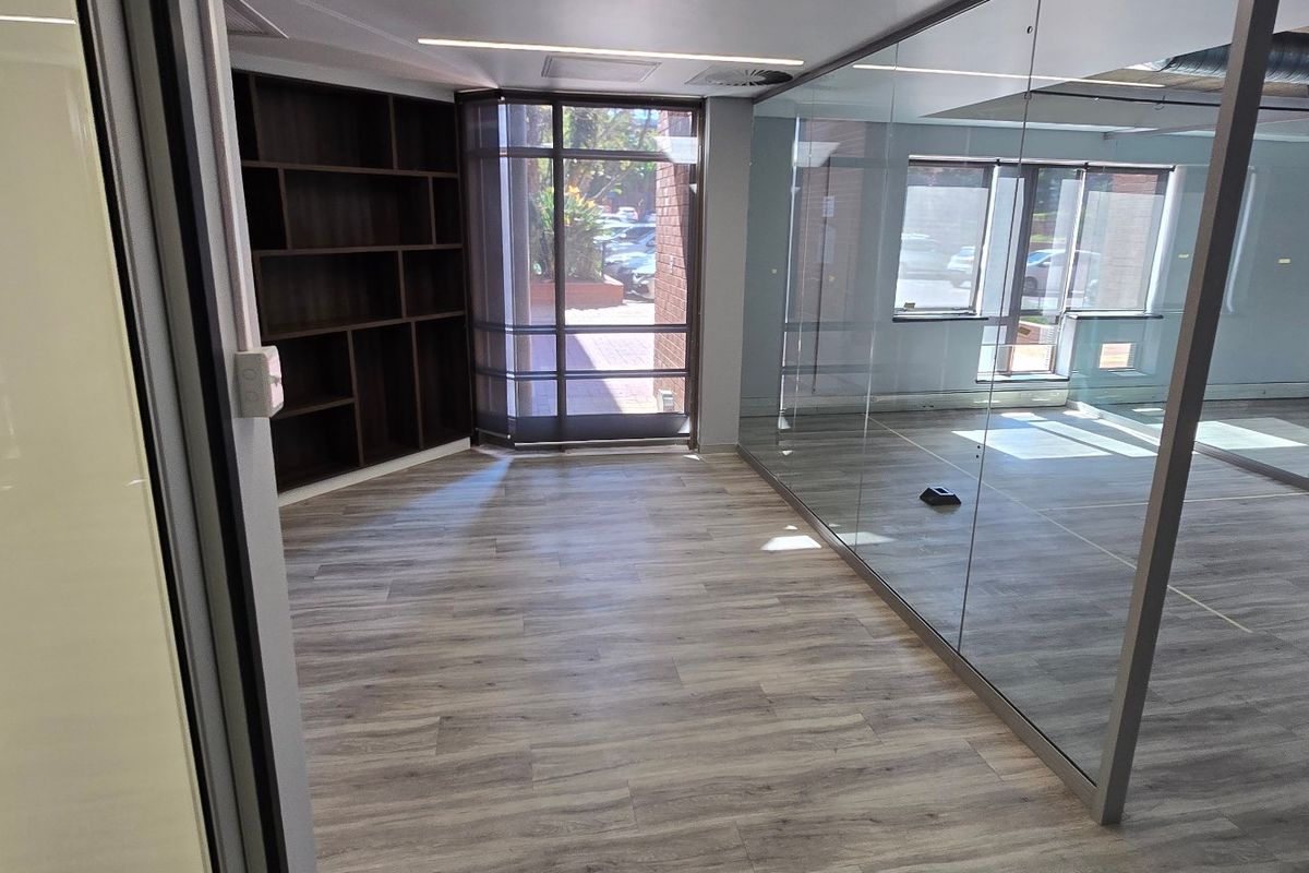 Modern commercial office space featuring glass partitioning, built-in shelving, laminate flooring, and exposed industrial-style ceiling ducting.