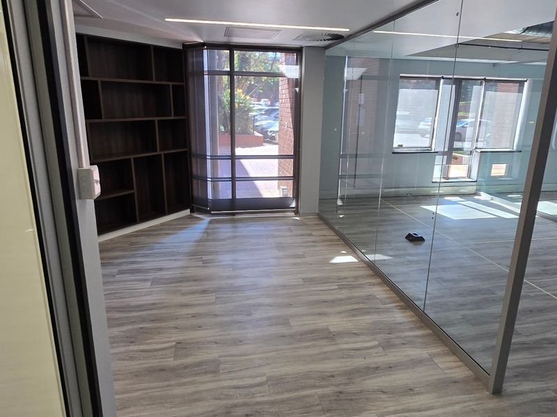 Modern commercial office space featuring glass partitioning, built-in shelving, laminate flooring, and exposed industrial-style ceiling ducting.