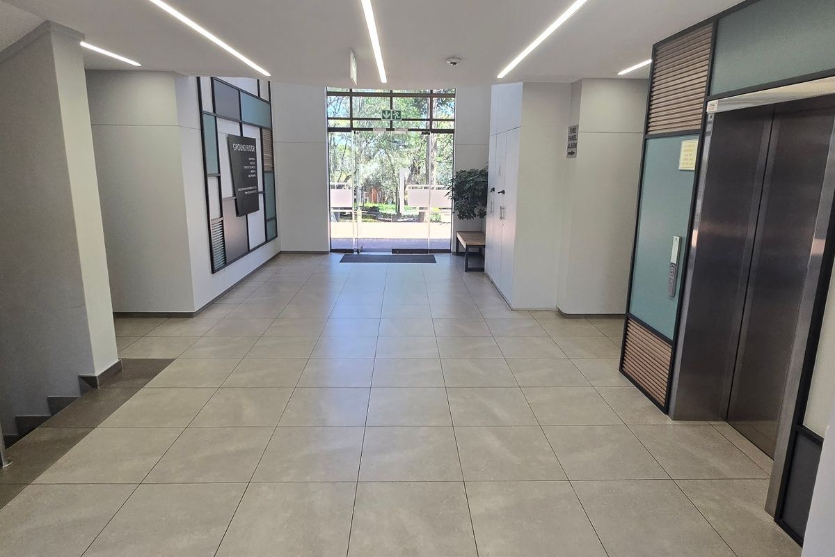 Modern commercial building lobby featuring tiled flooring, recessed linear lighting, a stainless steel elevator, and a glass entrance door leading to the exterior.