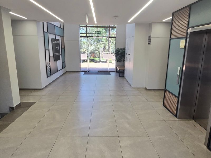 Modern commercial building lobby featuring tiled flooring, recessed linear lighting, a stainless steel elevator, and a glass entrance door leading to the exterior.