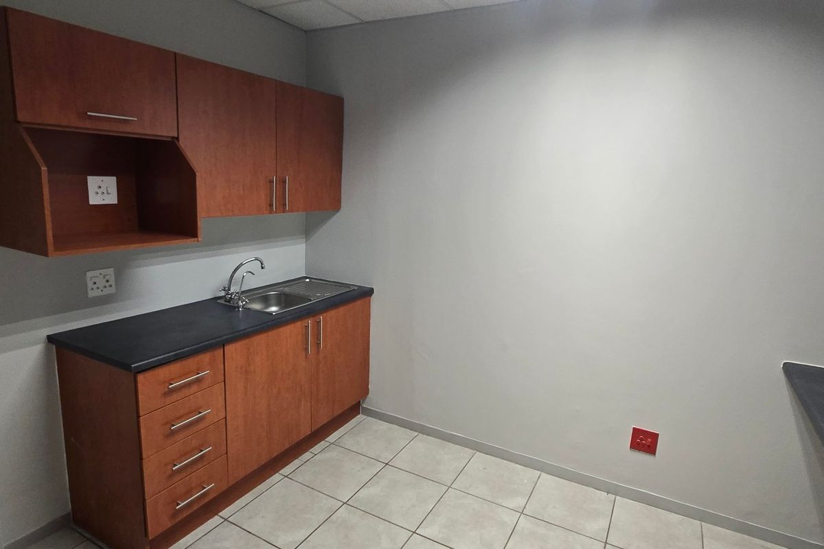 A compact commercial kitchenette featuring wood-finish cabinetry, a stainless steel sink, dark laminate countertops, and neutral tiled flooring.
