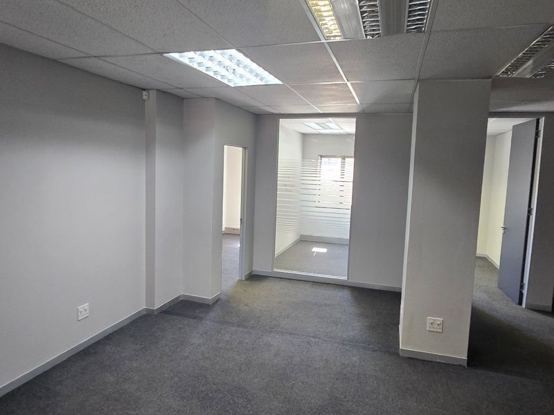 A vacant commercial office space featuring neutral grey carpeting, a suspended ceiling with recessed lighting, and a glass-partitioned private office area.