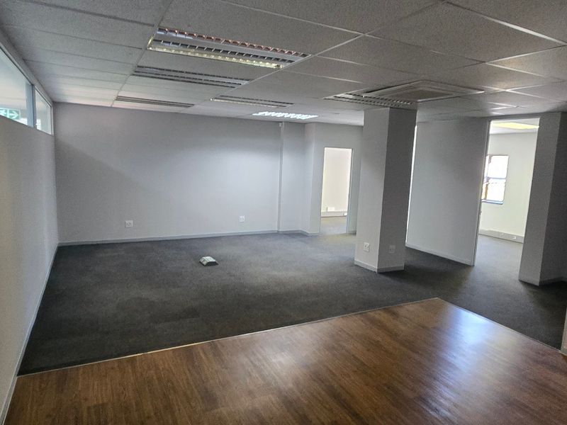 Open-plan commercial office space featuring a mix of carpet and laminate flooring, suspended ceilings with recessed lighting, and a central structural column.
