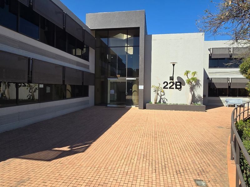 Modern commercial office building entrance featuring a paved courtyard, glass facade, and prominent building signage.