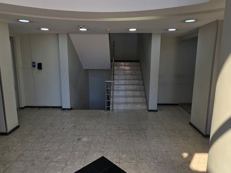 A spacious commercial building entrance lobby featuring tiled flooring, recessed ceiling lighting, and a central staircase with a metal handrail.