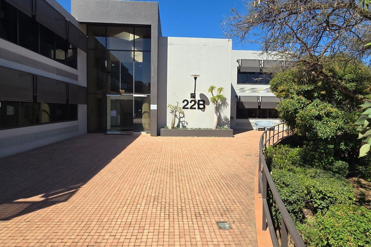 Front entrance view of a modern commercial office building featuring a paved courtyard, glass facade, and well-maintained landscaped garden areas.