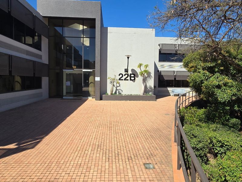 Front entrance view of a modern commercial office building featuring a paved courtyard, glass facade, and well-maintained landscaped garden areas.