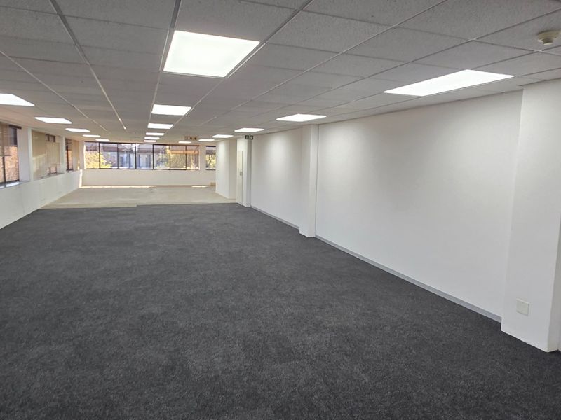 A spacious, open-plan commercial office area featuring neutral carpeting, recessed lighting, and large windows providing ample natural light.