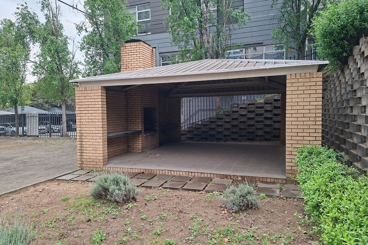 Covered outdoor patio area featuring a built-in brick braai, tiled flooring, and a metal roof, set against a landscaped retaining wall.