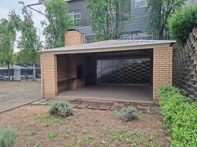 Covered outdoor patio area featuring a built-in brick braai, tiled flooring, and a metal roof, set against a landscaped retaining wall.