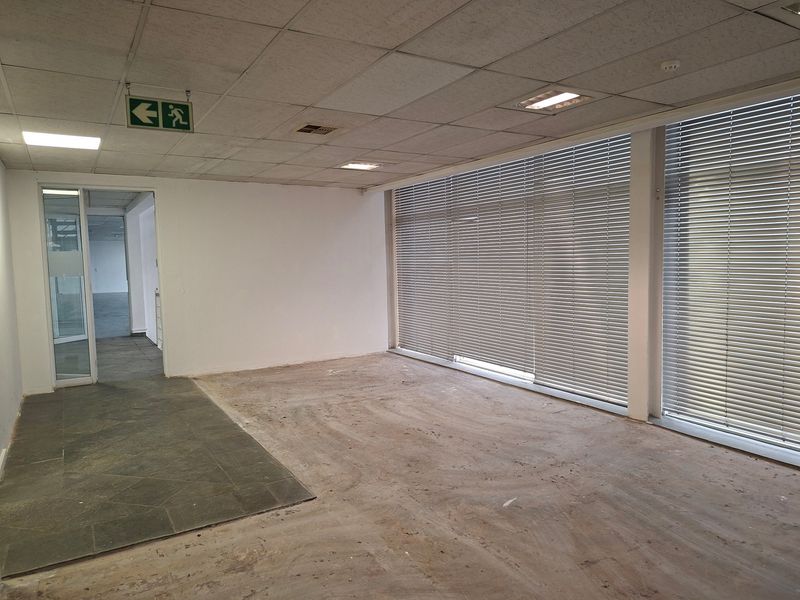 An empty industrial office space featuring large windows with venetian blinds, suspended ceilings with recessed lighting, and a glass partition door.