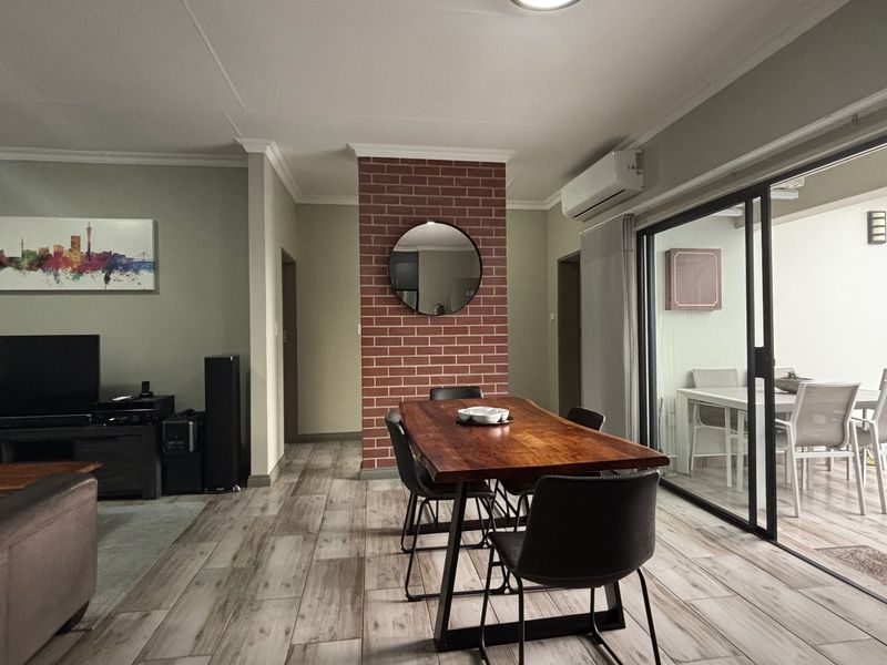 Spacious open-plan living and dining area featuring a brick accent wall, wood-look tile flooring, and seamless access to an outdoor patio through sliding glass doors.