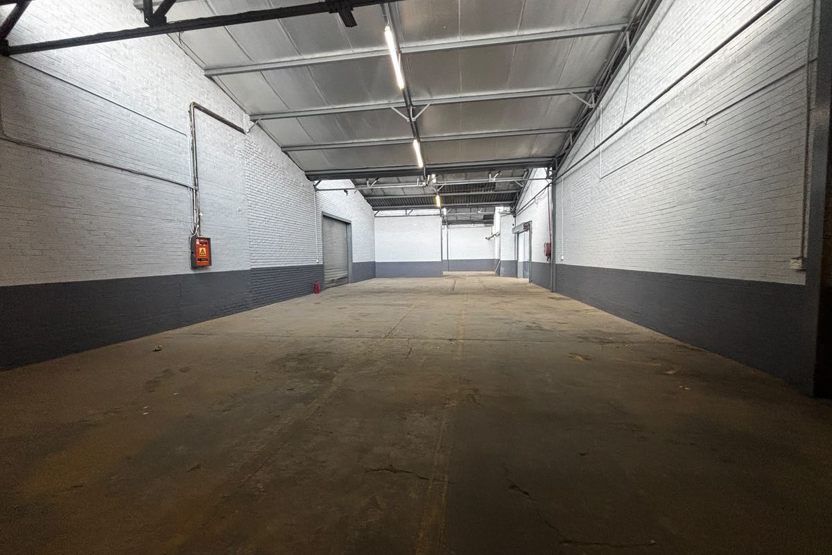 Spacious industrial warehouse unit featuring high ceilings with exposed steel trusses, painted brick walls, concrete flooring, and a roller shutter door.
