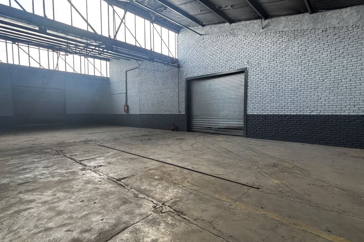 Spacious industrial warehouse unit featuring high ceilings with exposed steel trusses, concrete flooring, and a large roller shutter door for vehicle access.