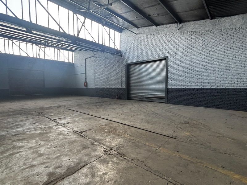 Spacious industrial warehouse unit featuring high ceilings with exposed steel trusses, concrete flooring, and a large roller shutter door for vehicle access.