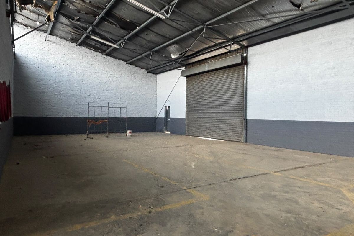 Spacious industrial warehouse unit featuring high ceilings with exposed steel trusses, concrete flooring, and a large roller shutter door for vehicle access.
