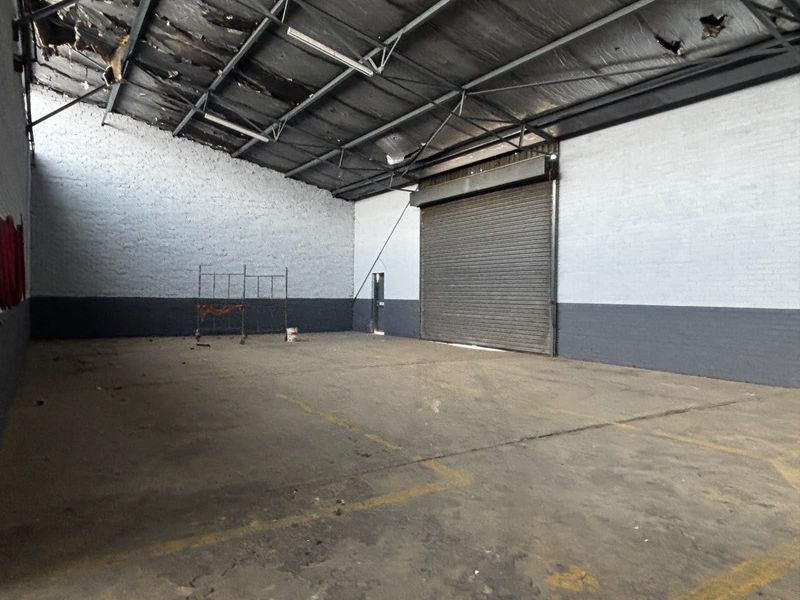 Spacious industrial warehouse unit featuring high ceilings with exposed steel trusses, concrete flooring, and a large roller shutter door for vehicle access.
