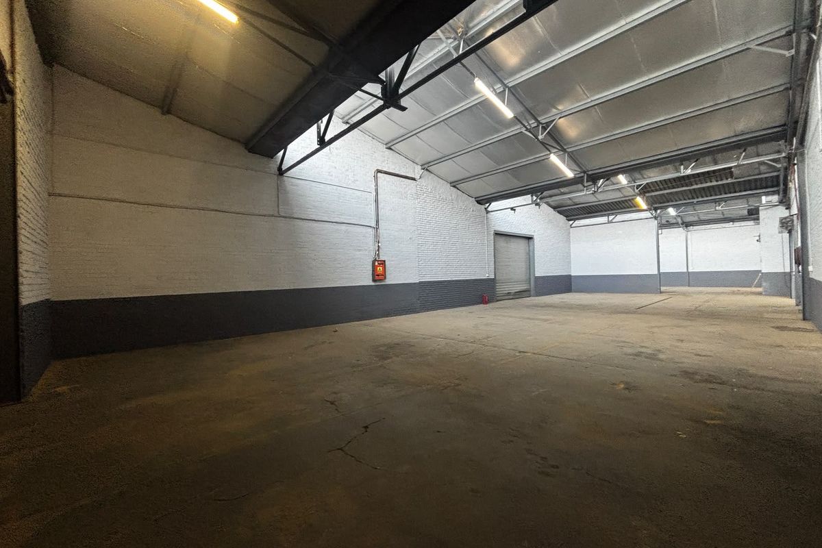 Spacious industrial warehouse unit featuring high ceilings with exposed steel trusses, concrete flooring, and a large roller shutter door for easy access.