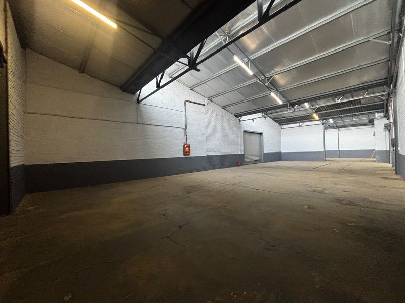 Spacious industrial warehouse unit featuring high ceilings with exposed steel trusses, concrete flooring, and a large roller shutter door for easy access.