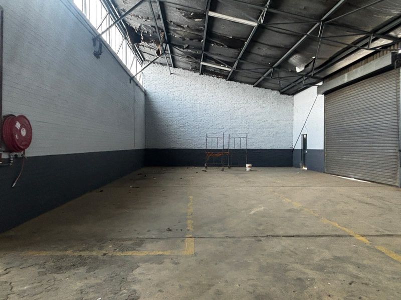 Spacious industrial warehouse unit featuring high ceilings with exposed steel trusses, concrete flooring, and a large roller shutter door for easy access.
