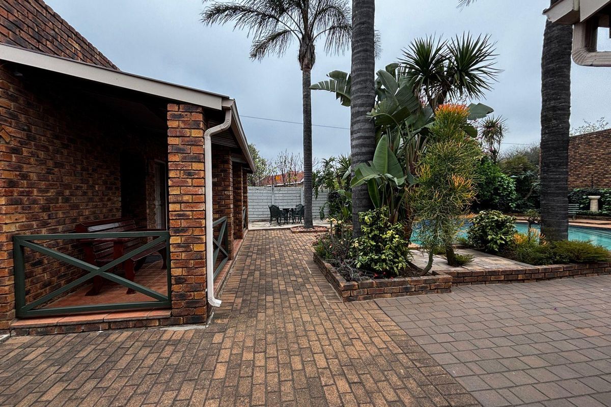 A rear exterior view featuring a brick-paved patio area, a covered veranda, established garden beds, and a swimming pool.