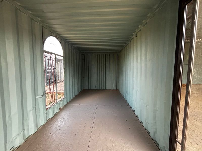 An empty industrial shipping container conversion space featuring corrugated steel walls, plywood flooring, and an arched window.