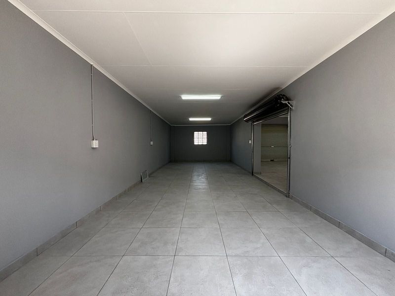 A clean, empty commercial utility space featuring grey tiled flooring, fluorescent lighting, and a secure roller shutter door entrance.