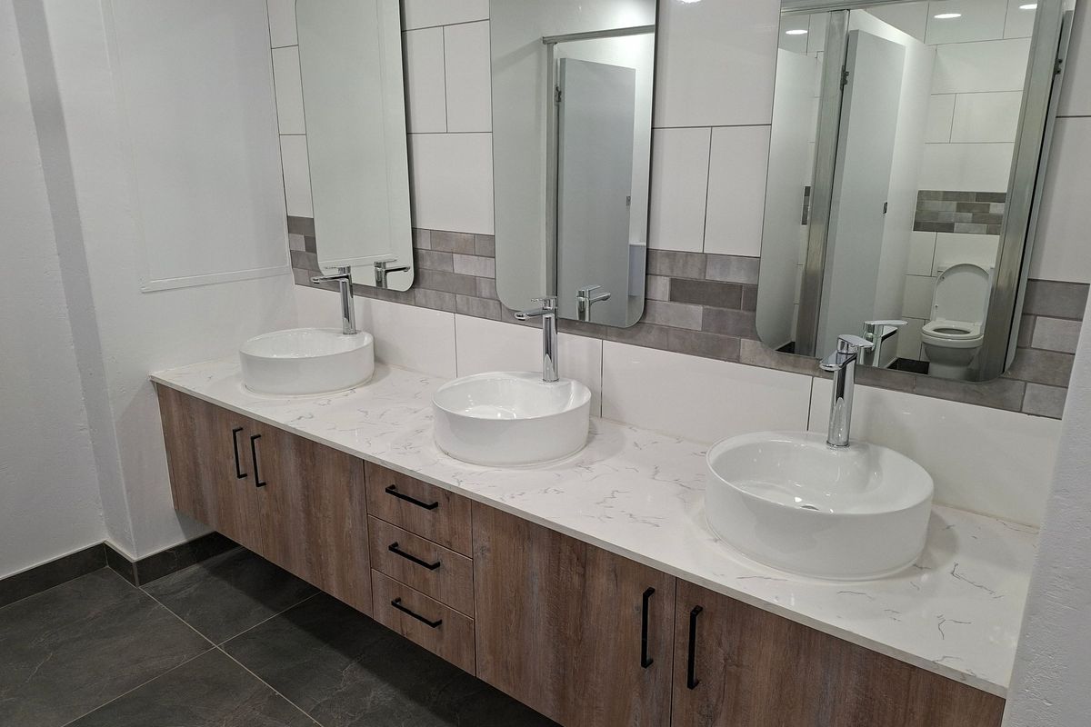 Modern commercial bathroom featuring a triple vanity with vessel sinks, stone countertop, wood-finish cabinetry, and tiled wall accents.