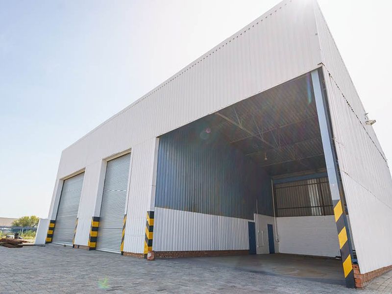 Industrial warehouse exterior featuring corrugated metal cladding, multiple roller shutter doors, and a paved driveway area.