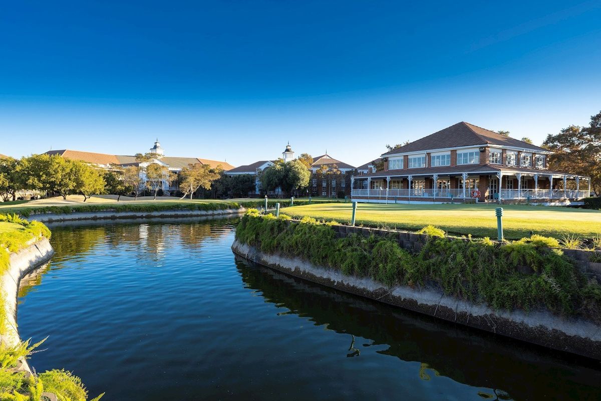 A scenic view of a commercial property featuring a well-maintained lawn, a water feature, and a spacious covered veranda overlooking the grounds.