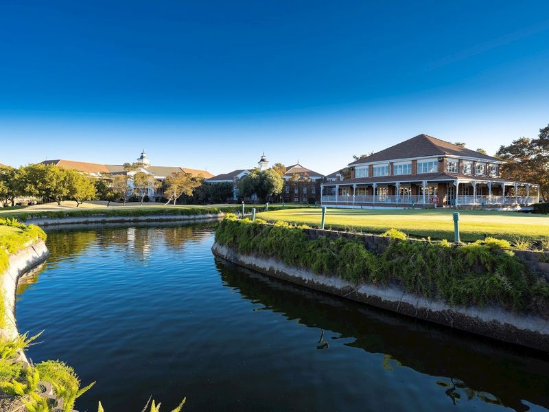 A scenic view of a commercial property featuring a well-maintained lawn, a water feature, and a spacious covered veranda overlooking the grounds.