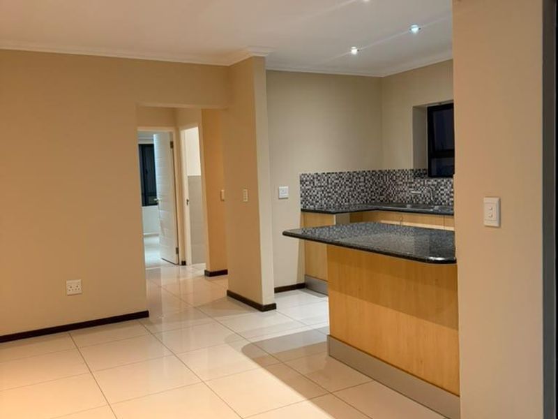 An open-plan living area featuring a kitchen with granite countertops, wooden cabinetry, and a breakfast bar, finished with neutral walls and tiled flooring.